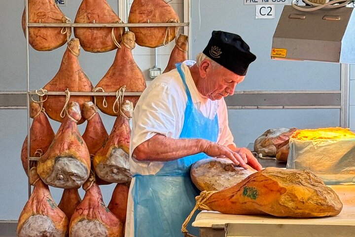 Prosciutto di Parma producer trimming a curing ham by hand, with hams hanging on the rack behind him